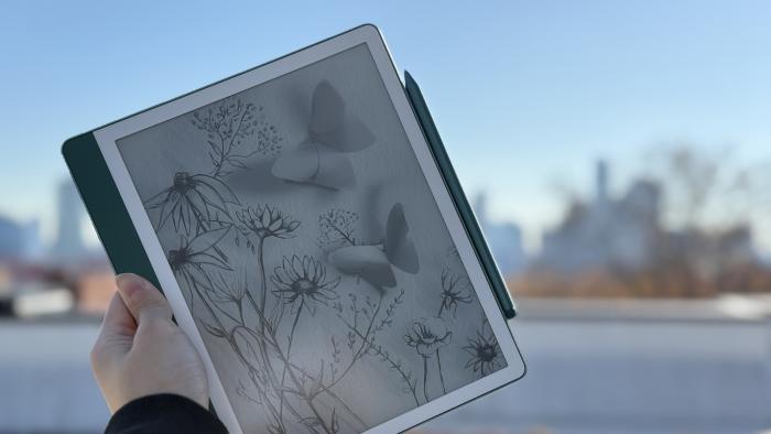 The Amazon Kindle Scribe 2nd gen being held up in mid air, with a Premium Pen attached to its right side. In the background is the NYC skyline.