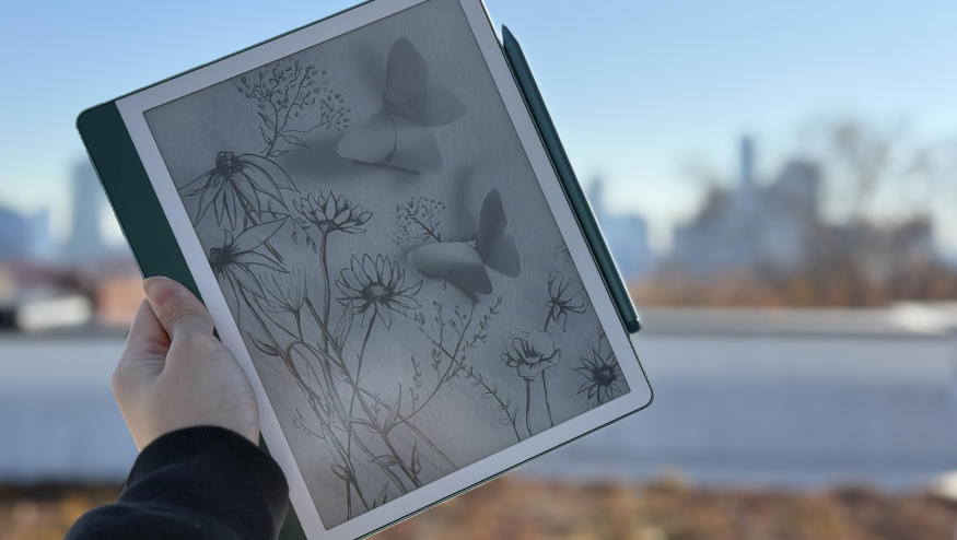 The Amazon Kindle Scribe 2nd gen being held up in mid air, with a Premium Pen attached to its right side. In the background is the NYC skyline.