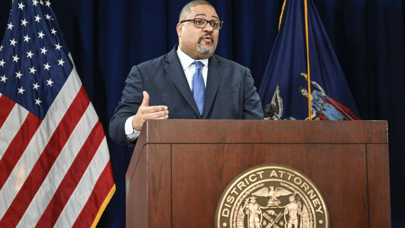 A person in a suit behind a wooden podium with the American flag in the background.